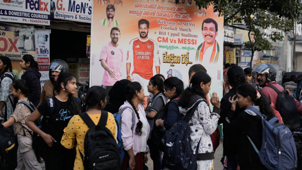 College students stands in front of a billboard of Argentine soccer star Lionel Messi and Telangana Chief Minister Anumula Revanth Reddy, hours before Messi's arrival in the capital city Hyderabad, India, Saturday, Dec. 13, 2025. (AP Photo/Mahesh Kumar A.) MKA103
