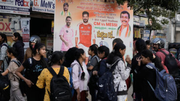 College students stands in front of a billboard of Argentine soccer star Lionel Messi and Telangana Chief Minister Anumula Revanth Reddy, hours before Messi's arrival in the capital city Hyderabad, India, Saturday, Dec. 13, 2025. (AP Photo/Mahesh Kumar A.)  MKA103